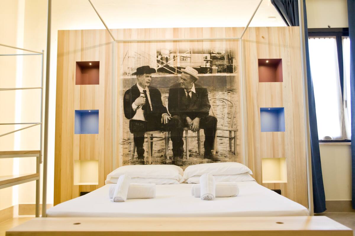 A hotel room with a large bed, two bedside tables, and a wall-mounted artwork of two men sitting on a bench.