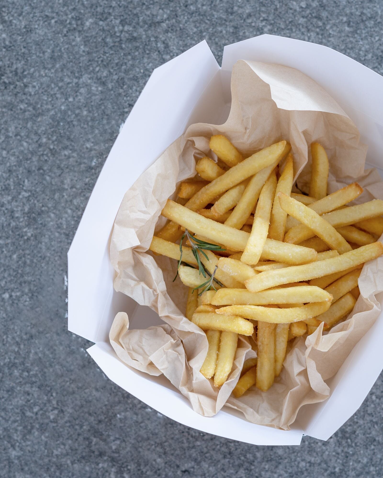 Un récipient rempli de frites, surmonté d’une branche de romarin, posé sur une surface texturée.