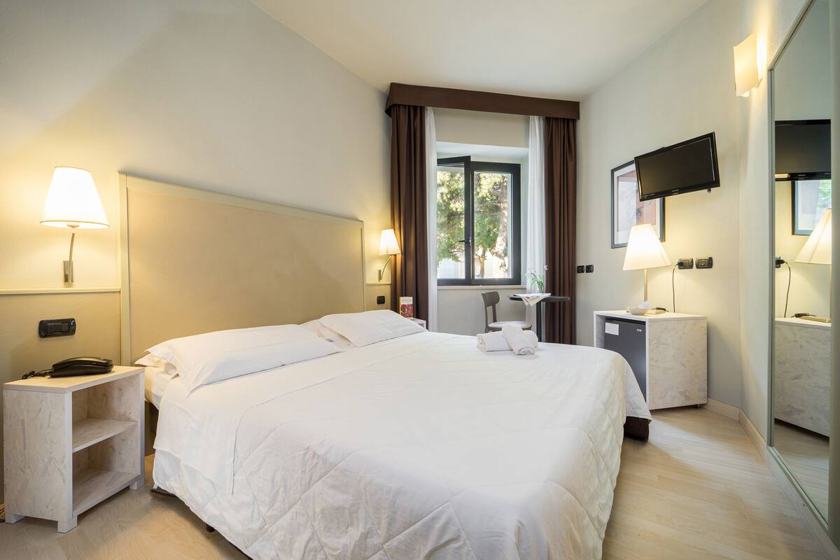 A hotel room with a large bed, bedside tables with lamps, a desk with a chair, a TV mounted on the wall, and a window with curtains.