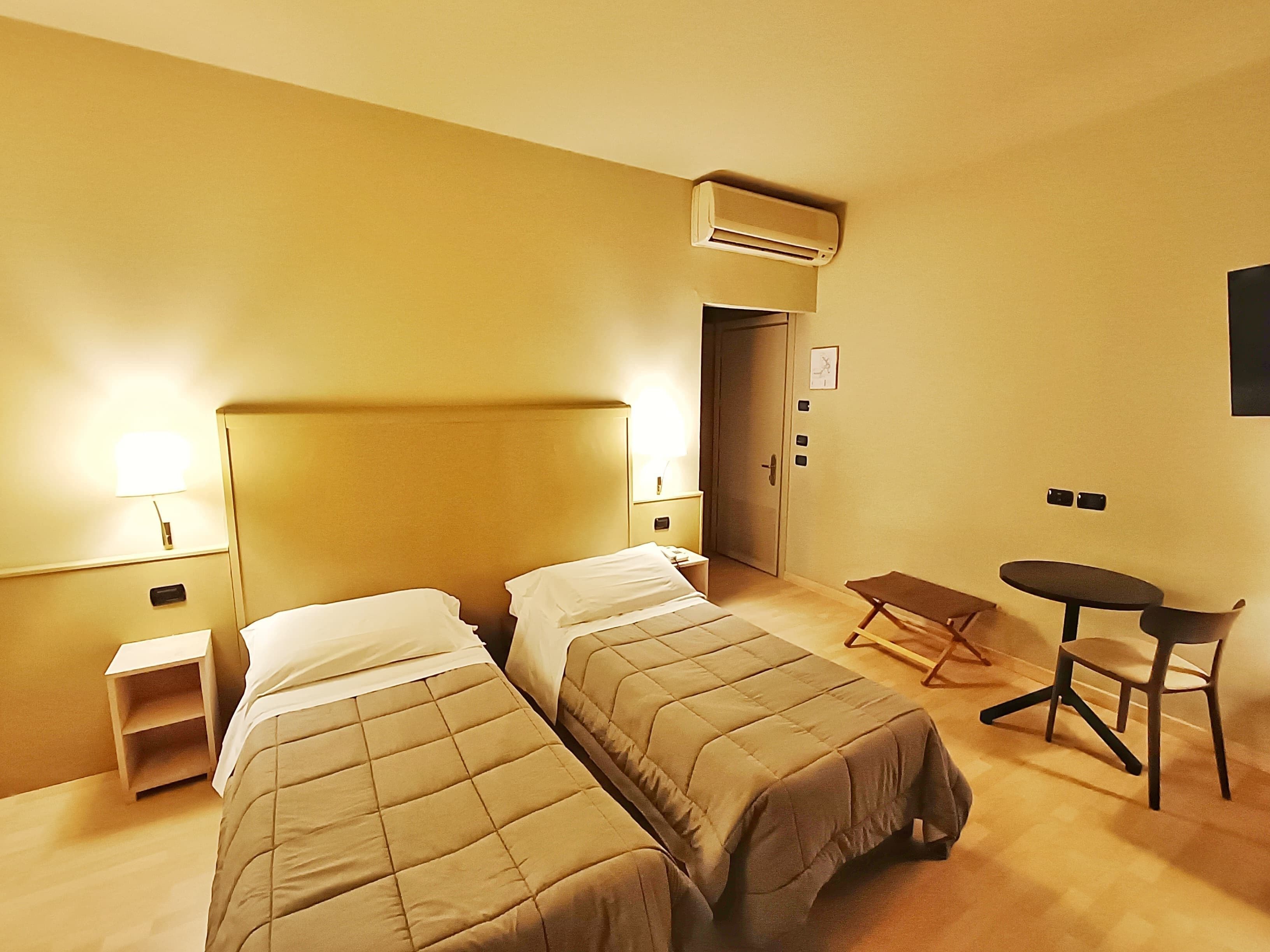 Ein Hotelzimmer mit zwei Betten, einem kleinen Tisch, einem Stuhl, einer Klimaanlage, einer Nachttischlampe und einer Tür.