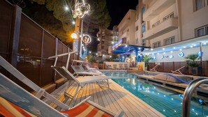 Un área de piscina con sillones de descanso, una terraza de madera y un edificio con un letrero al fondo.