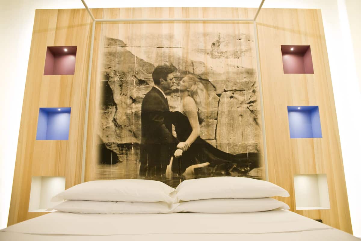 A bedroom with a large bed, a wall-mounted painting of a couple, and two wall sconces.
