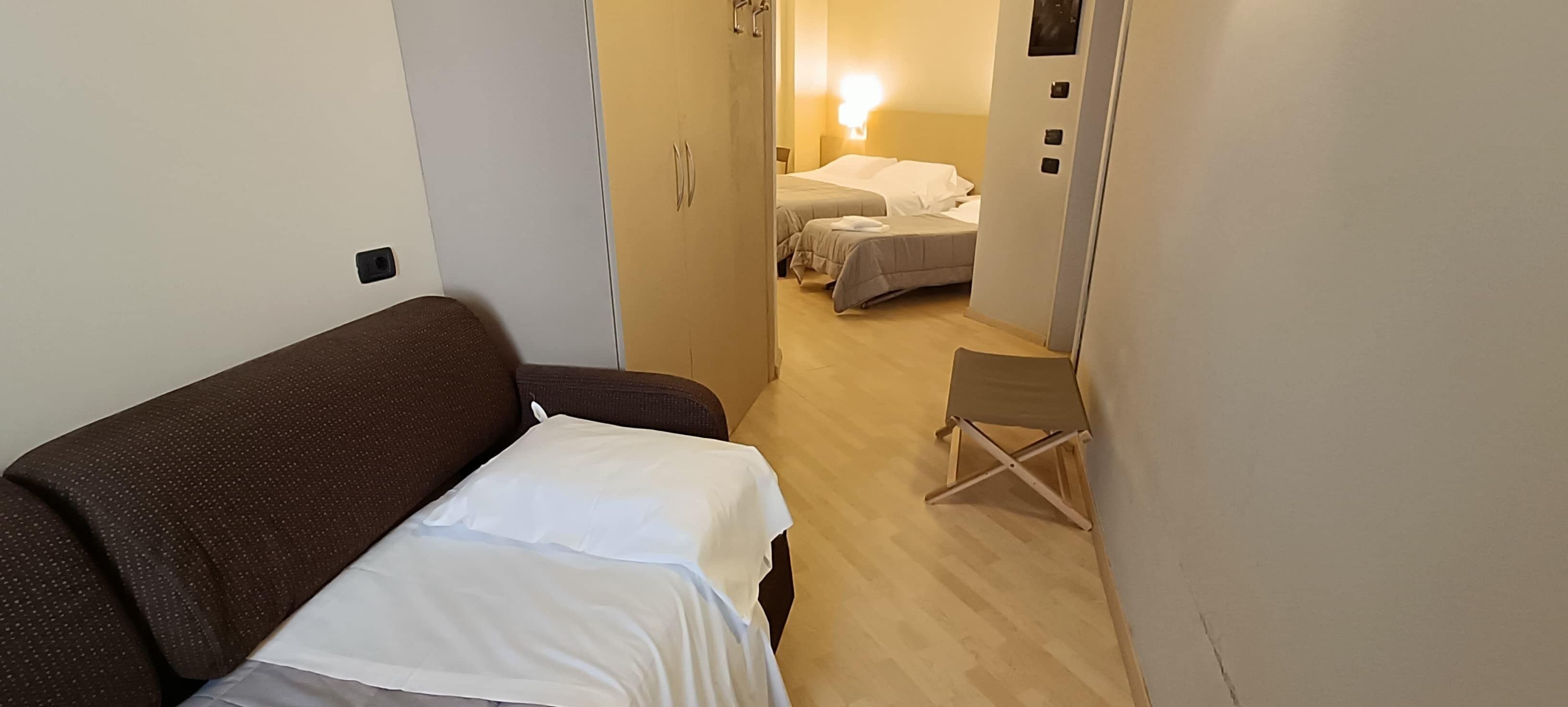 Quarto de hotel com um sofá marrom, uma cadeira dobrável pequena e duas camas com roupa de cama branca.
