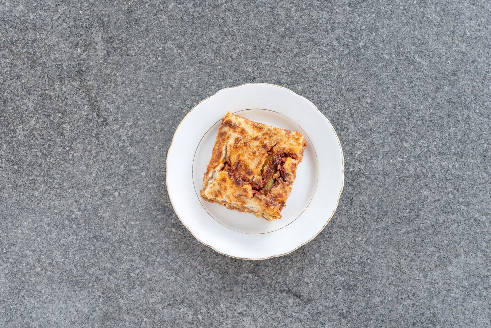 En enda skiva lasagne på en vit tallrik med guldkant, placerad på en grå, strukturerad yta.