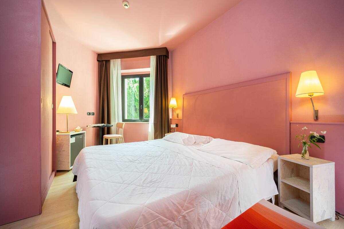 A hotel room with a large bed, bedside lamps, a TV, a desk, and a window with curtains.