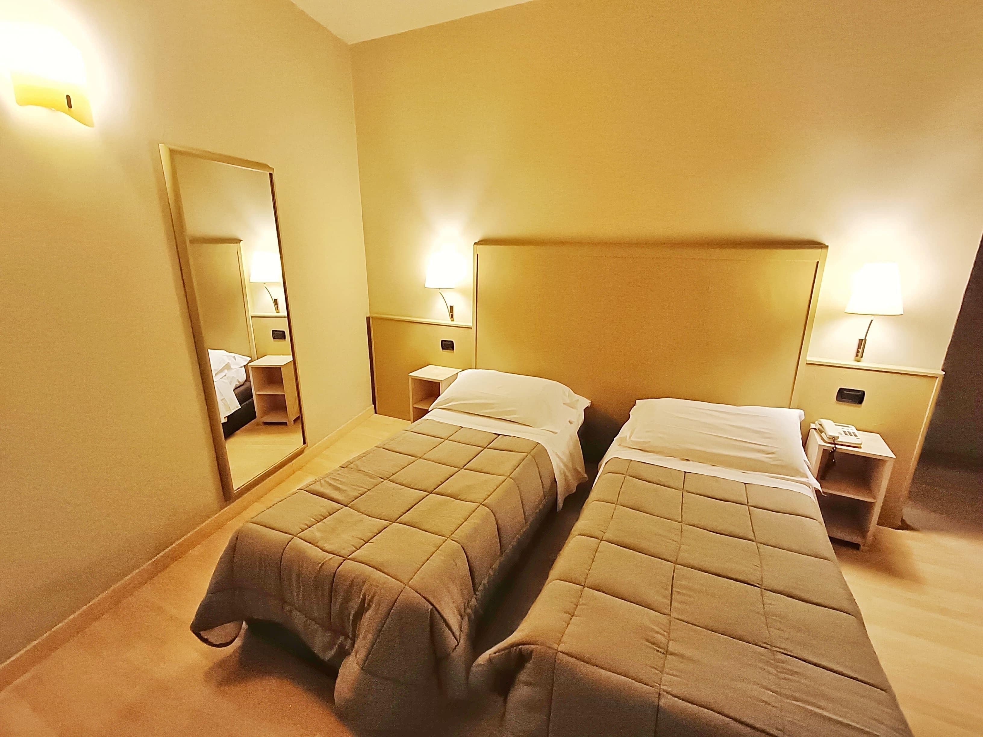 Ein Hotelzimmer mit zwei Betten, Nachttischlampen, einem Nachttisch mit Telefon und einem Spiegel an der Wand.