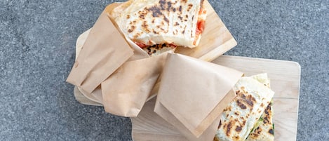 Ein Holzschnittbrett mit drei in braunes Papier gewickelten Quesadillas, auf einer grauen, strukturierten Oberfläche platziert.