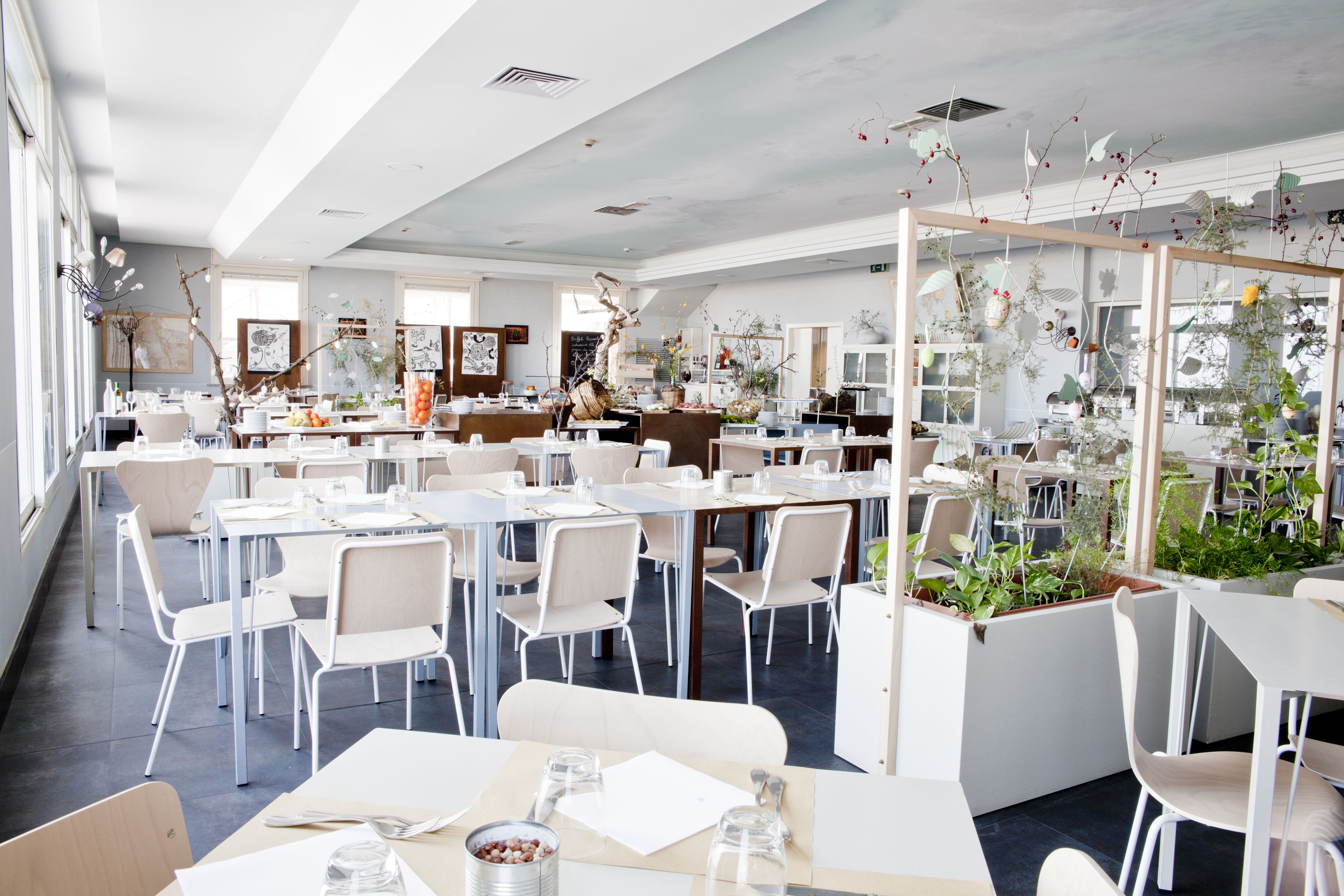 A spacious dining area with multiple tables and chairs, large windows, and decorative plants.