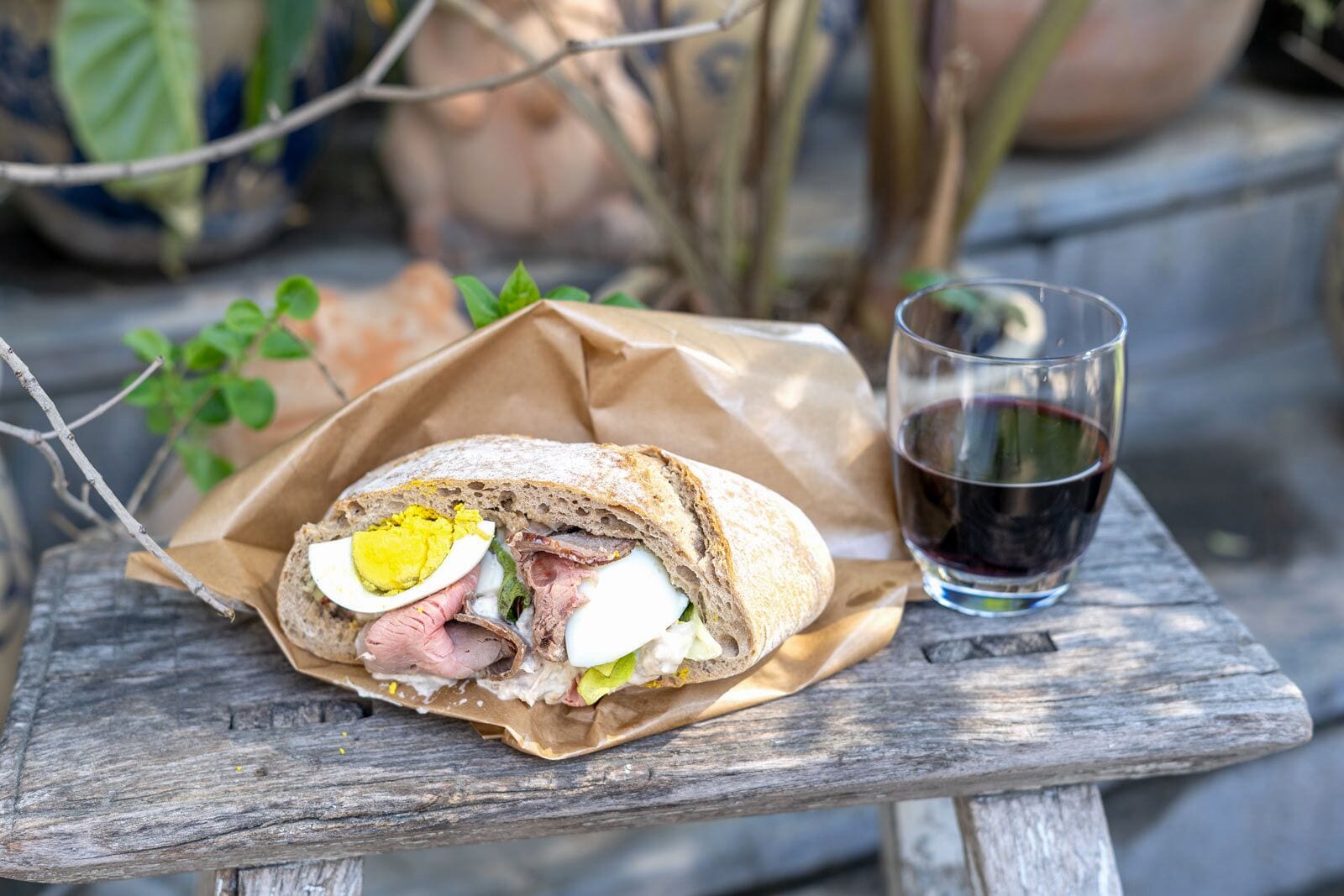 A sandwich on a wooden table with a glass of red wine beside it.