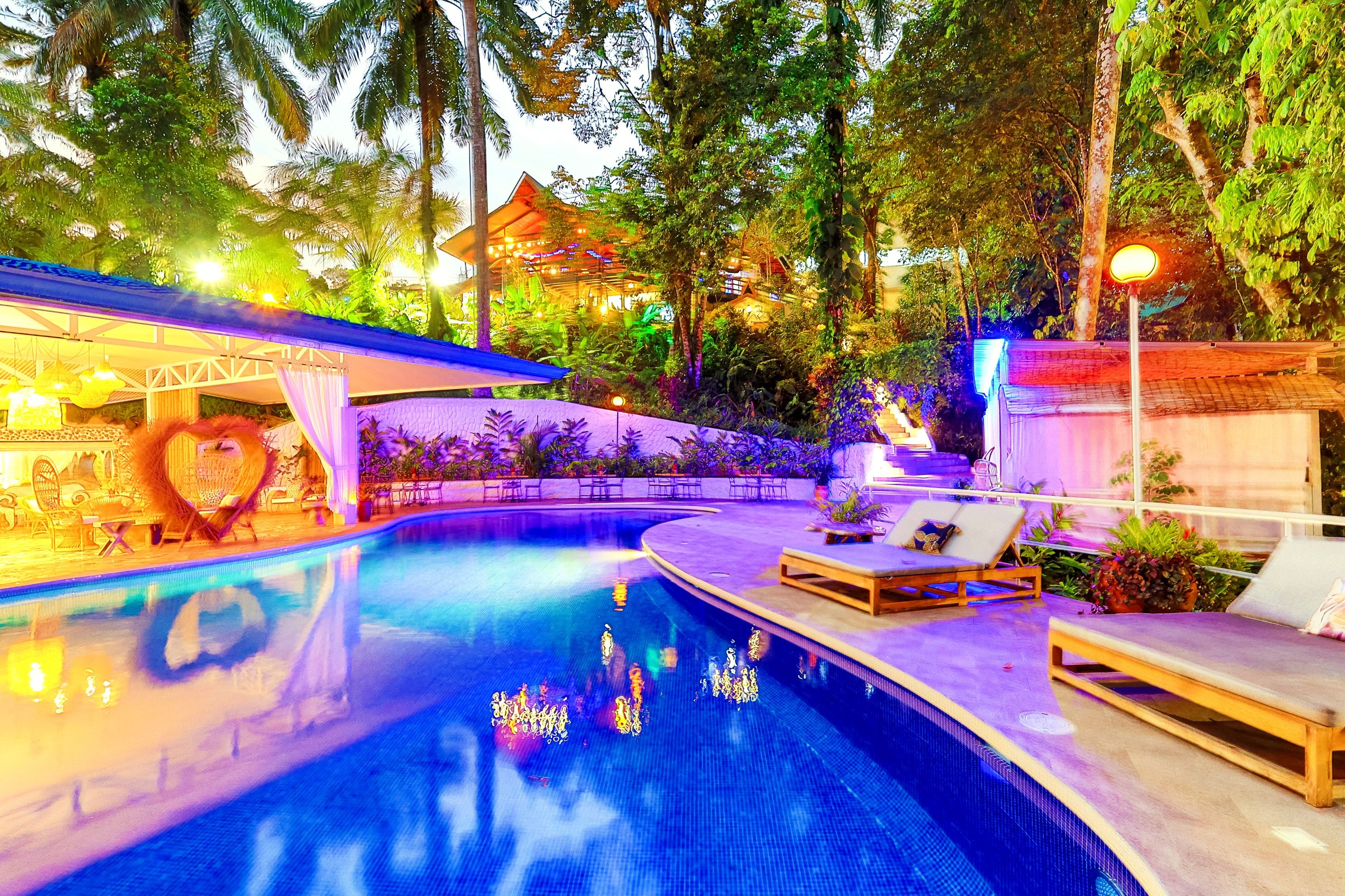 A swimming pool with lounge chairs and a covered seating area, surrounded by lush greenery and a building with a traditional roof.