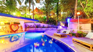 A swimming pool with lounge chairs and a covered seating area, surrounded by lush greenery and a building with a traditional roof.
