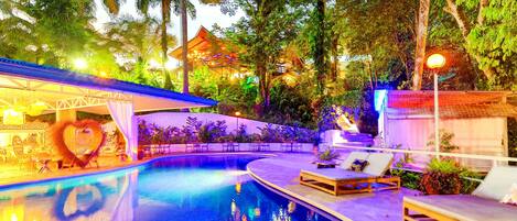 A swimming pool with lounge chairs and a covered seating area, surrounded by lush greenery and a building with a traditional roof.