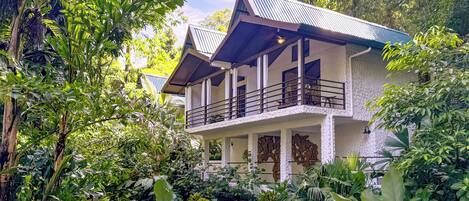 A two-story house with a balcony, surrounded by lush greenery.