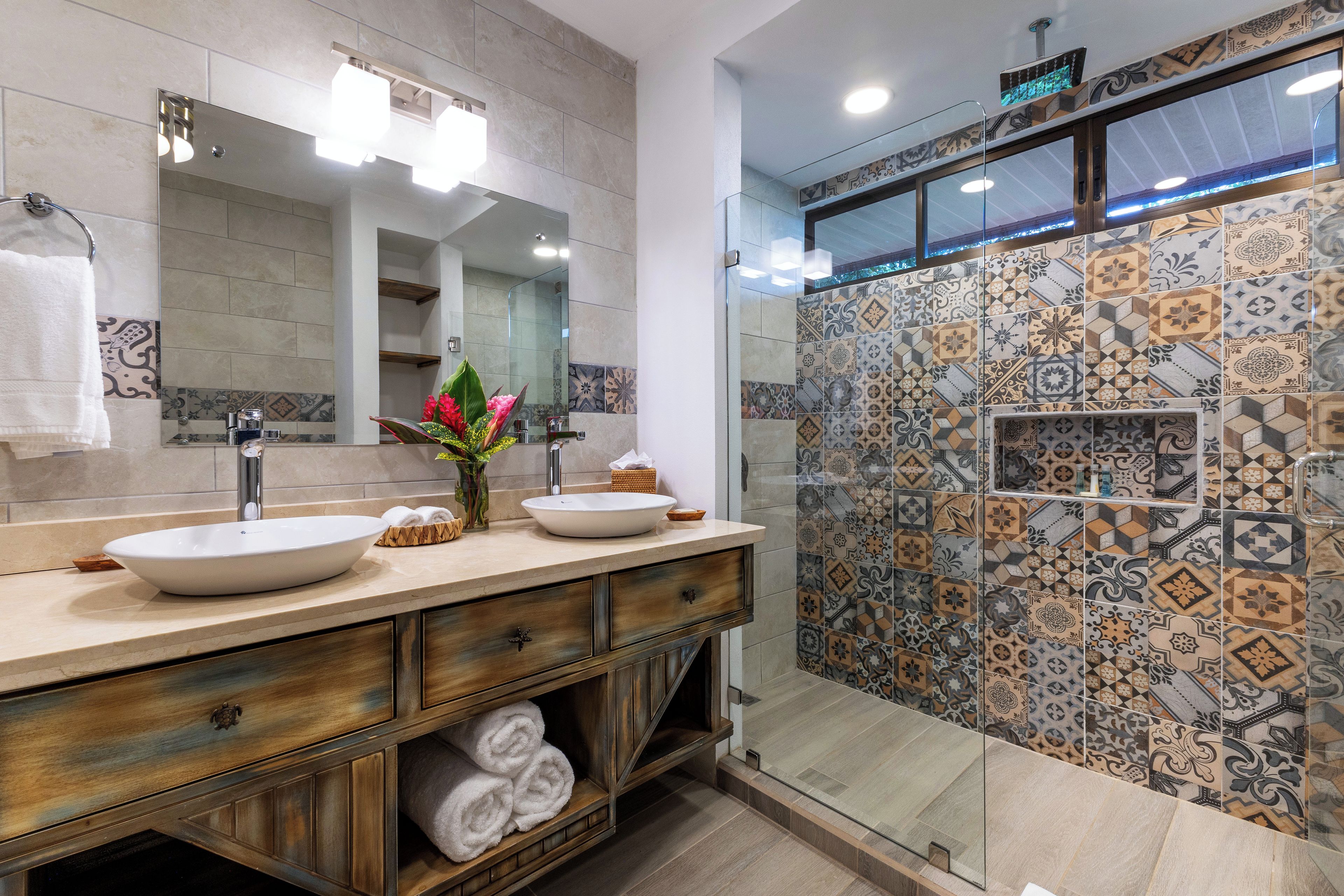 Une salle de bain moderne avec un grand miroir, deux vasques, une douche aux carreaux décoratifs et un meuble-lavabo en bois surmonté de serviettes.