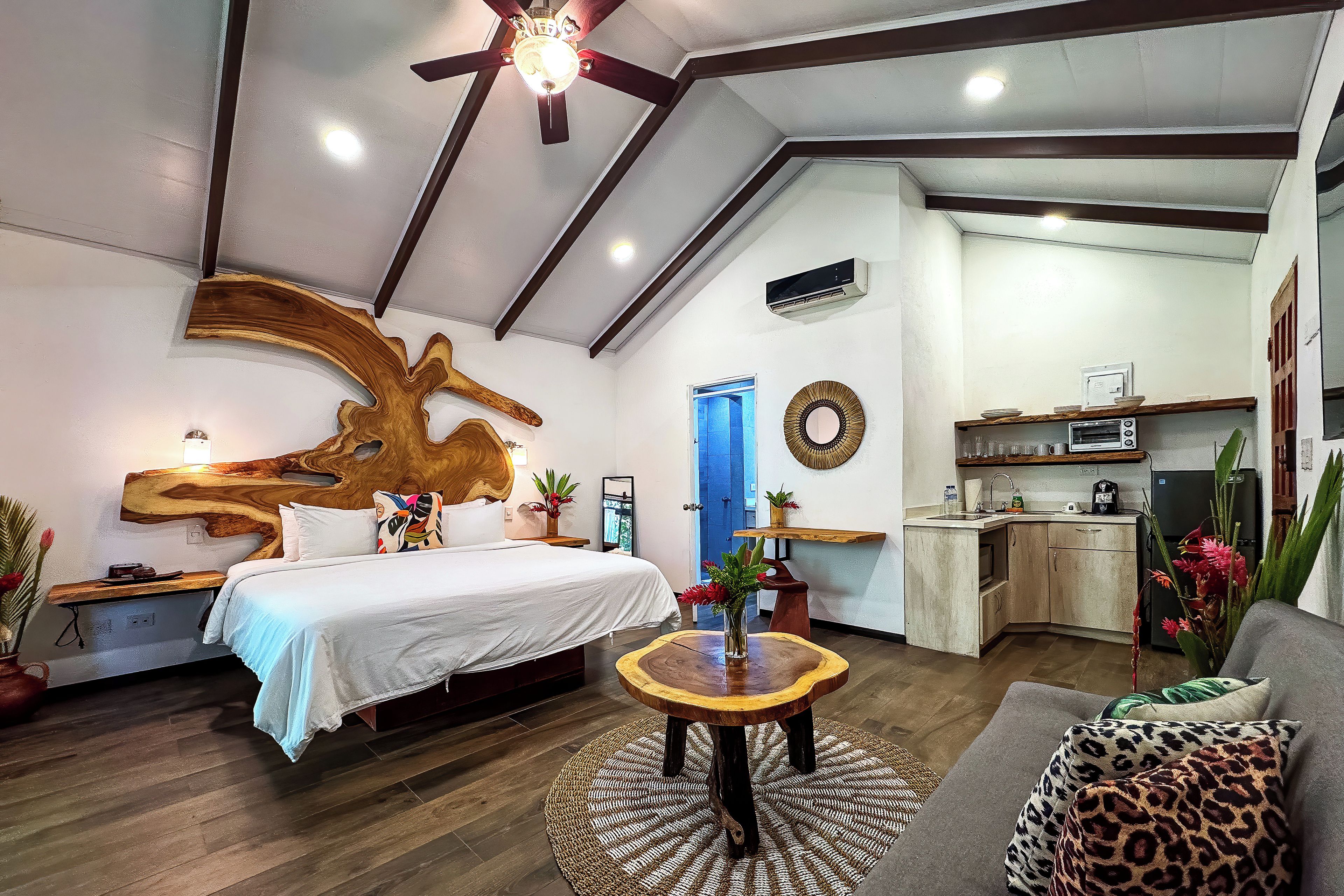 A modern bedroom with a large bed, a wooden headboard, a small round table with a vase, and a kitchenette in the background.
