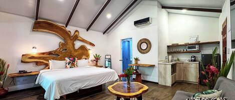 A modern bedroom with a large bed, a wooden headboard, a small round table with a vase, and a kitchenette in the background.