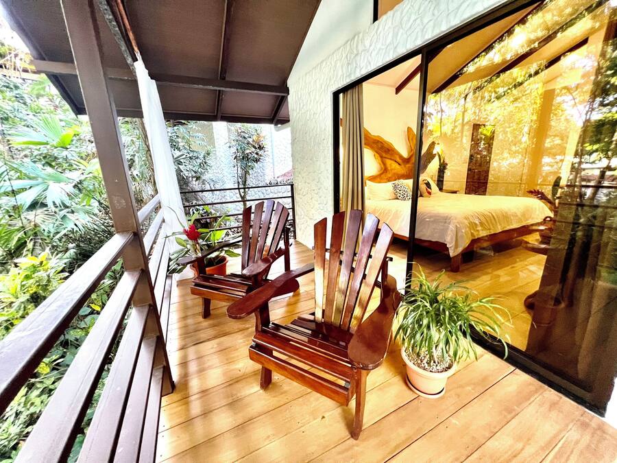 Ein Balkon mit Holzstühlen und Blumentöpfen, der auf ein Schlafzimmer mit Bett und großem Fenster blickt.