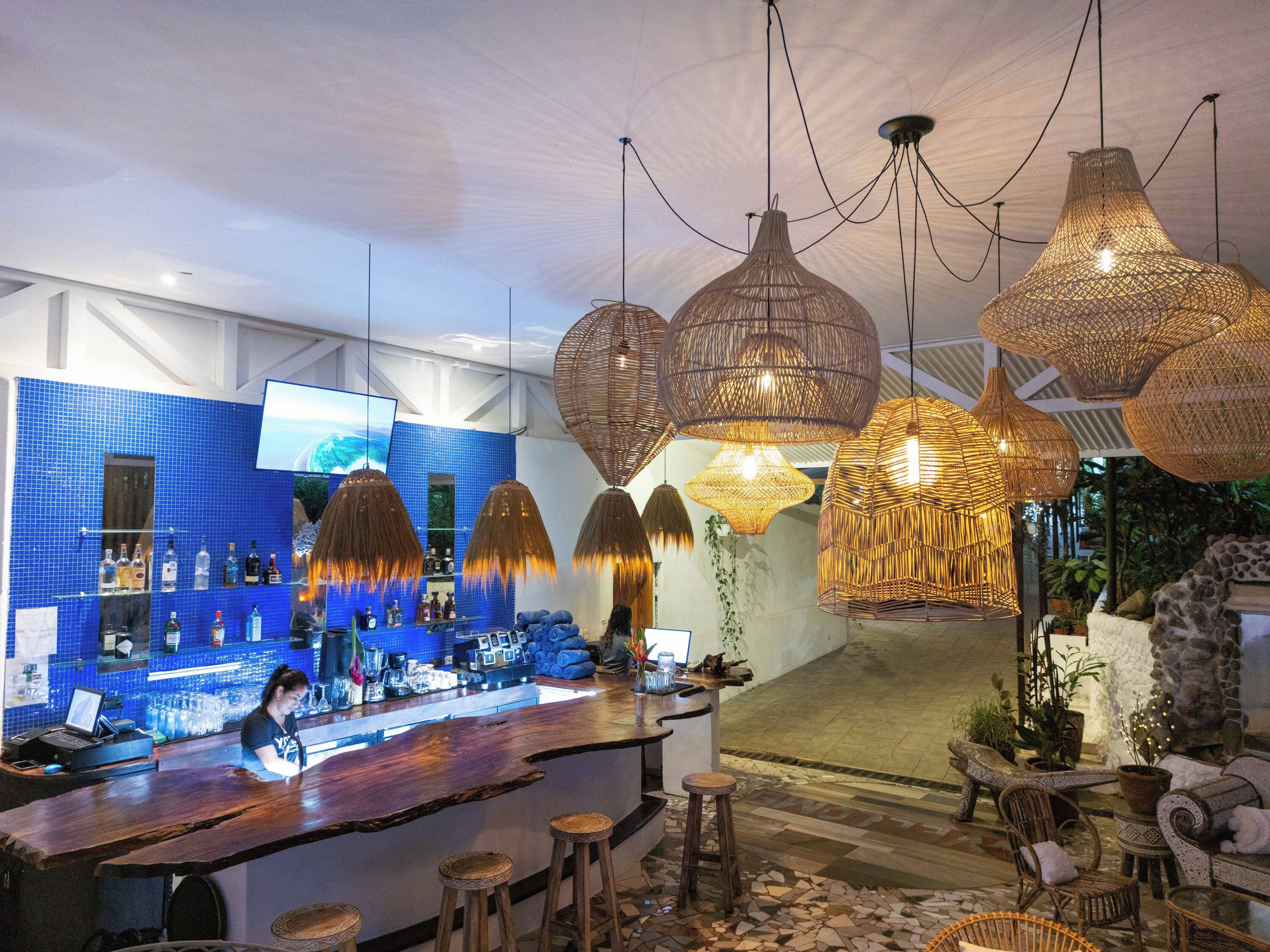 A bar area with a blue tiled backsplash, hanging rattan chandeliers, and a bar counter with stools.