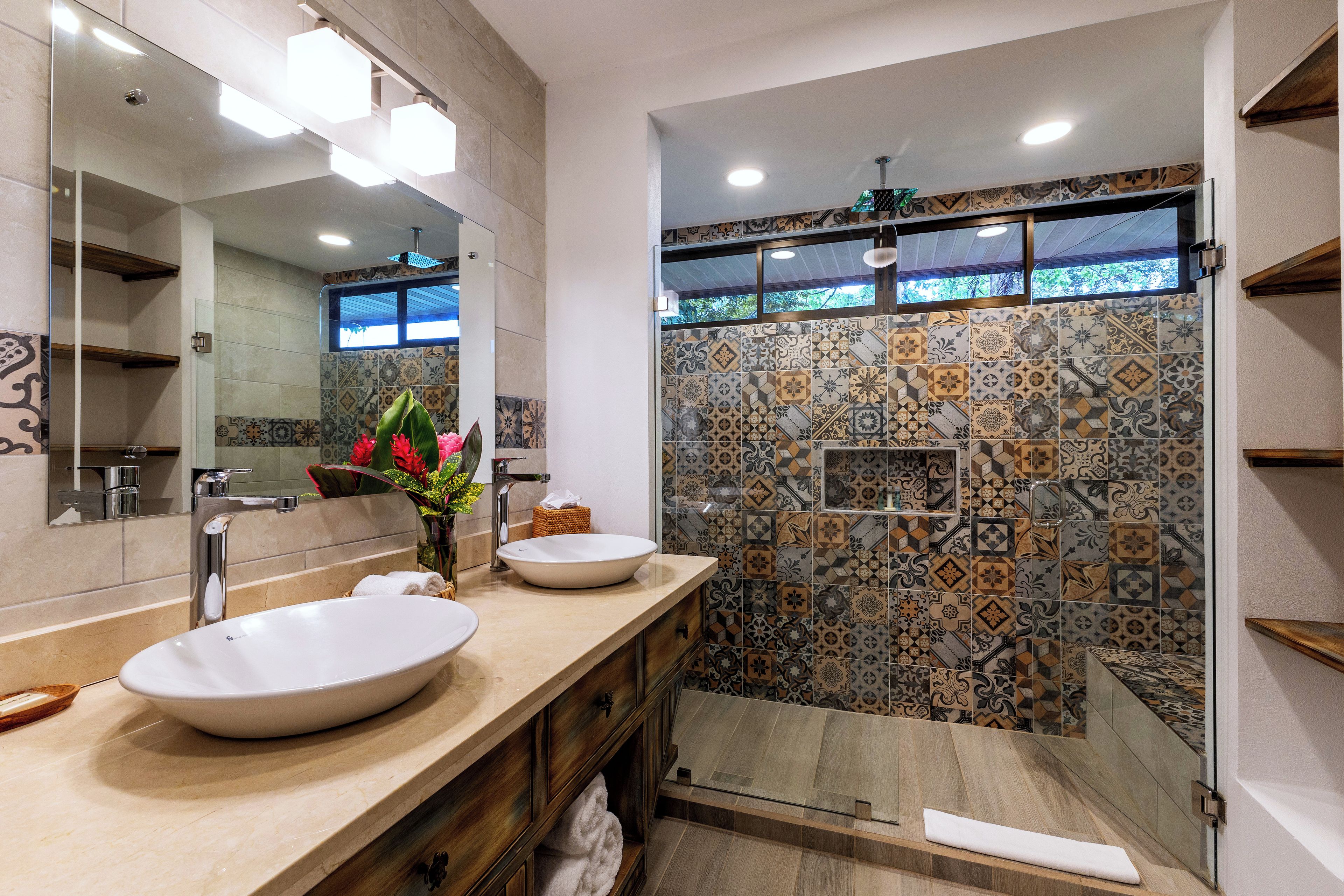 A modern bathroom with a large mirror, two sinks, a shower with patterned tiles, and a wooden vanity.