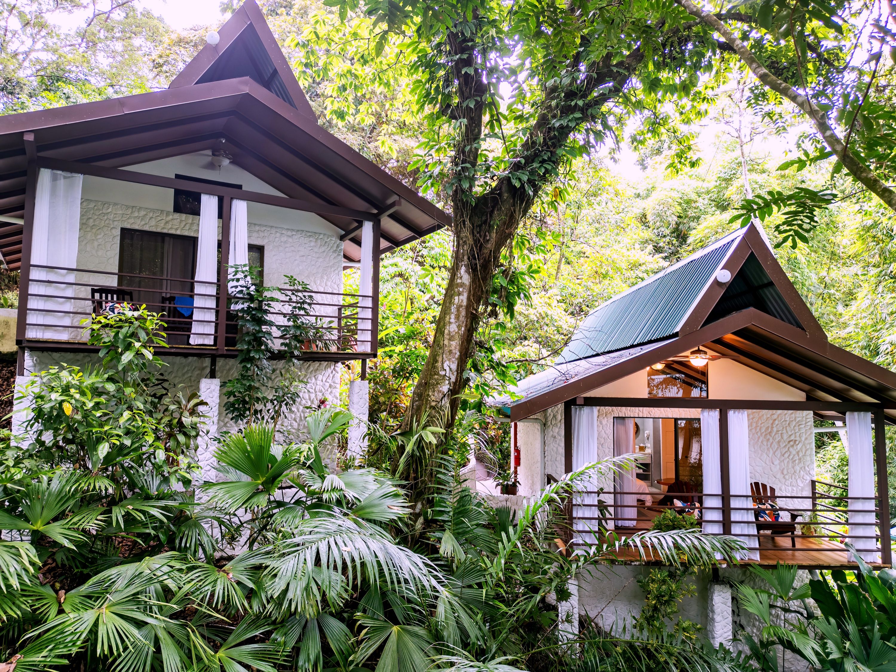 A two-story house with a balcony and a covered porch, surrounded by lush greenery.