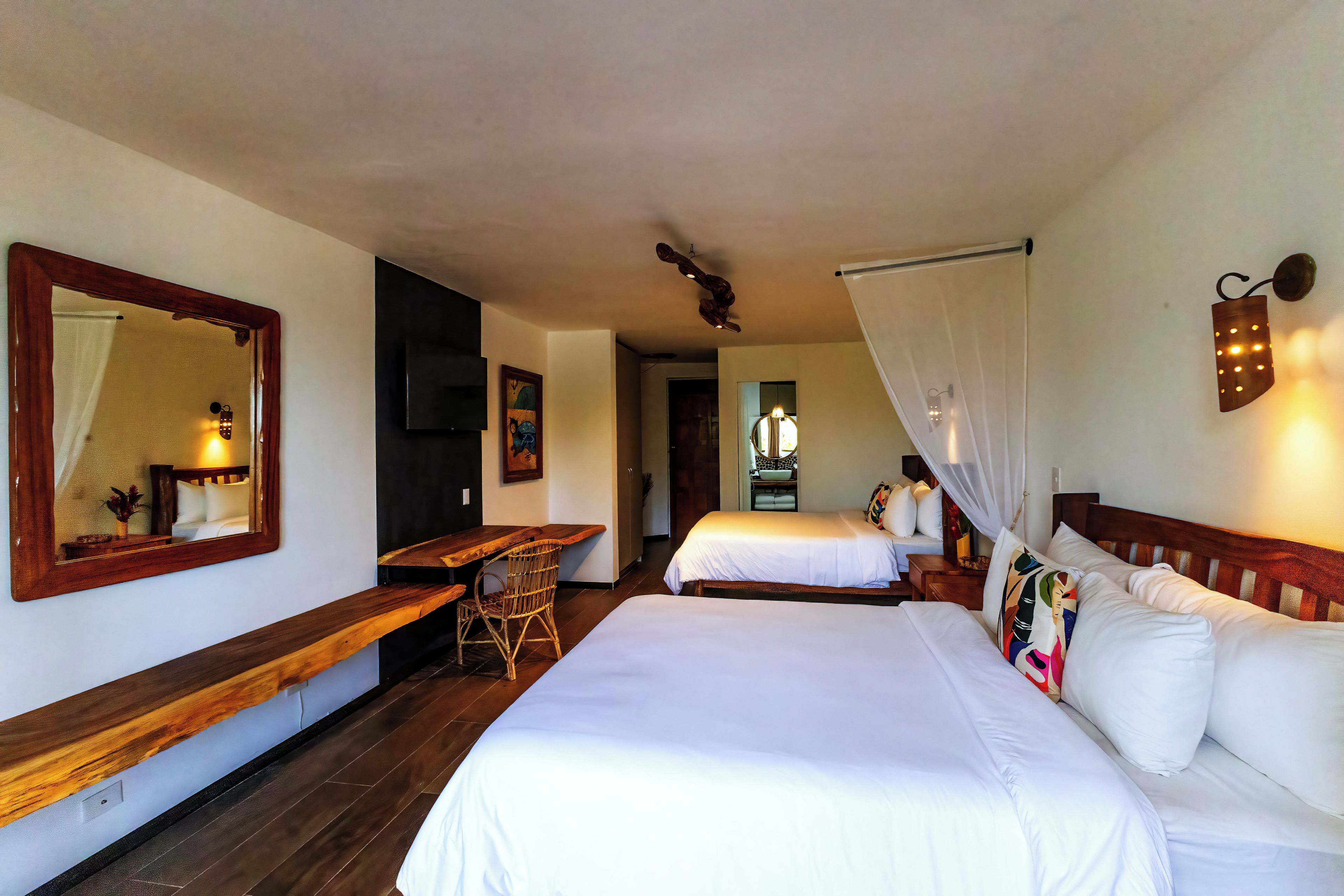 A hotel room with a large bed, a smaller bed, a desk, a chair, a mirror, and a wall-mounted light fixture.