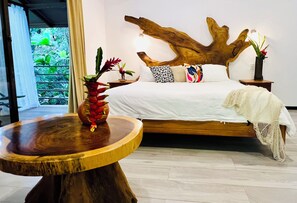 A bedroom with a large wooden headboard, a bed with white linens, a bedside table with a vase of flowers, and a view of greenery through a window. - ECOTICA Boutique Hotel & Spa. (Quepos)