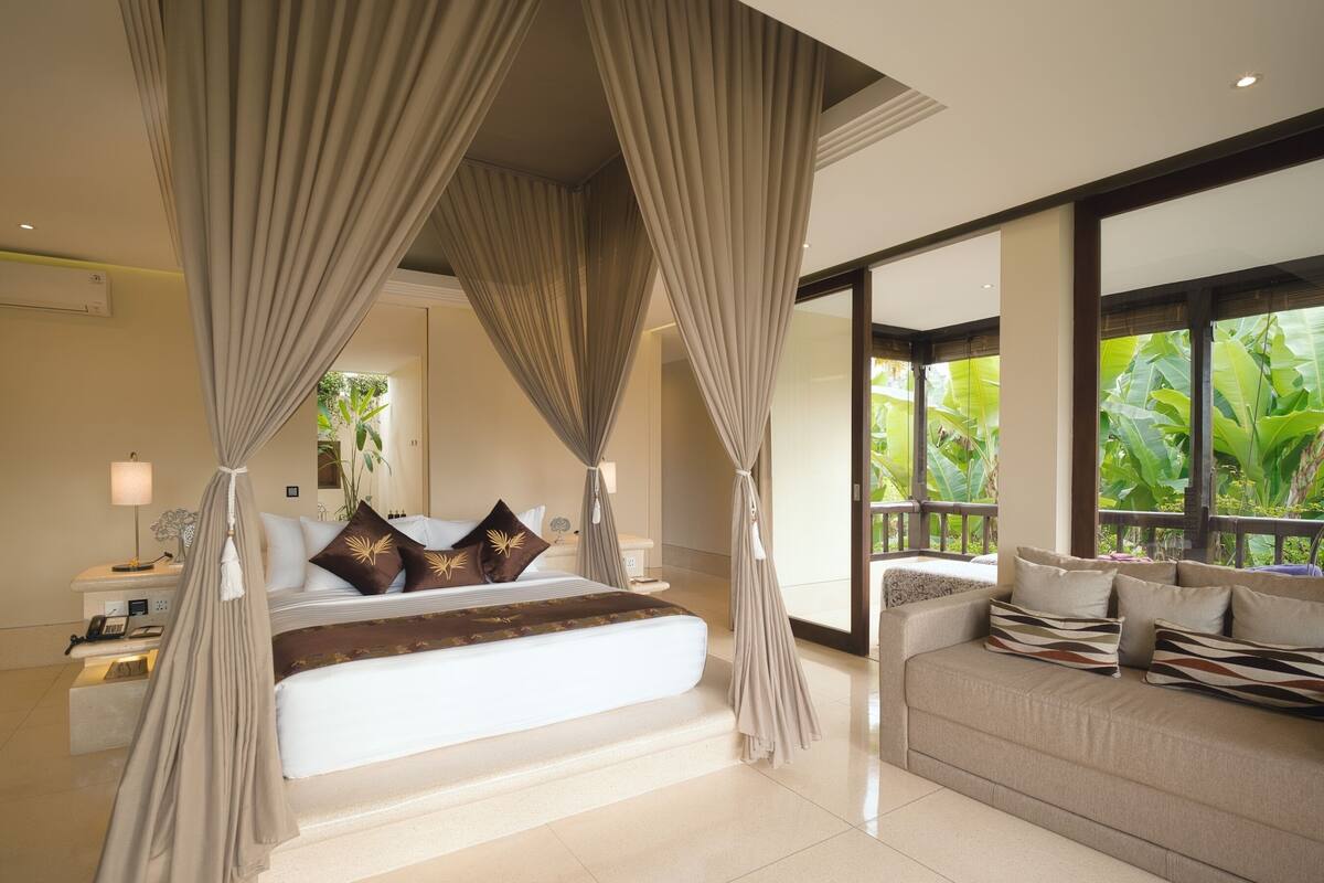 A bedroom with a bed, a sofa, curtains, and a view of greenery.