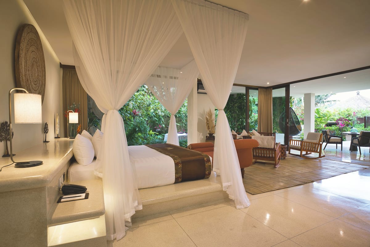 A bedroom with a canopy bed, a bedside table with a lamp, and a view of the garden.