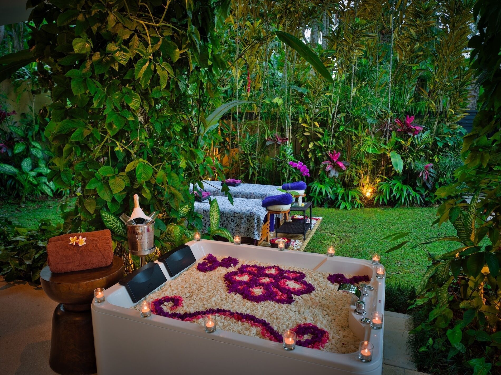 Un área de spa con una bañera llena de arroz y pétalos de flores, rodeada de exuberante follaje y velas.