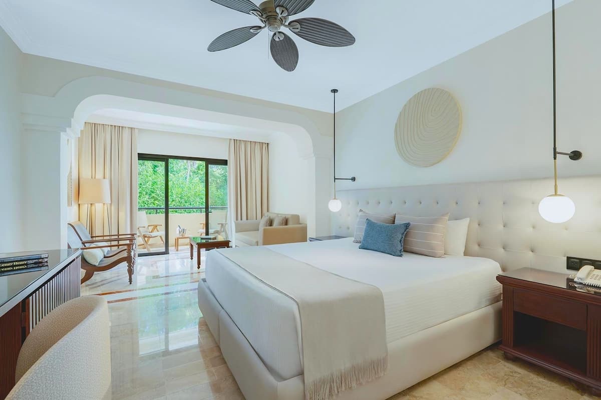 A spacious bedroom with a large bed, a desk, and a chair. There is a balcony with curtains and a view of greenery.