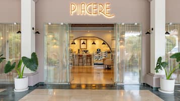 Entrada de hotel com fachada de vidro, uma placa com a palavra 'PIACERE' e uma área de recepção com assentos e balcão.