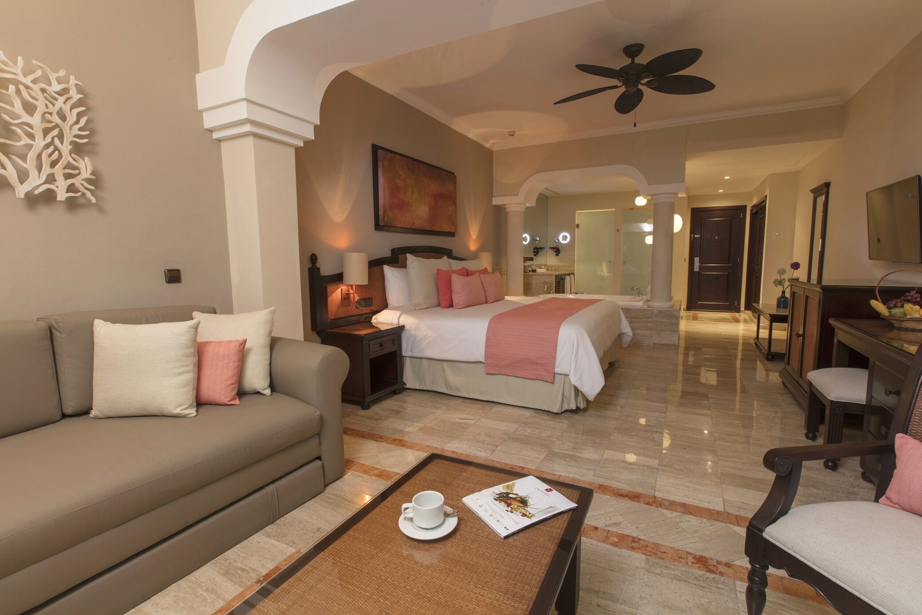 A hotel room with a sofa, a bed, a desk, a chair, a coffee table, a ceiling fan, and a bathroom visible in the background.