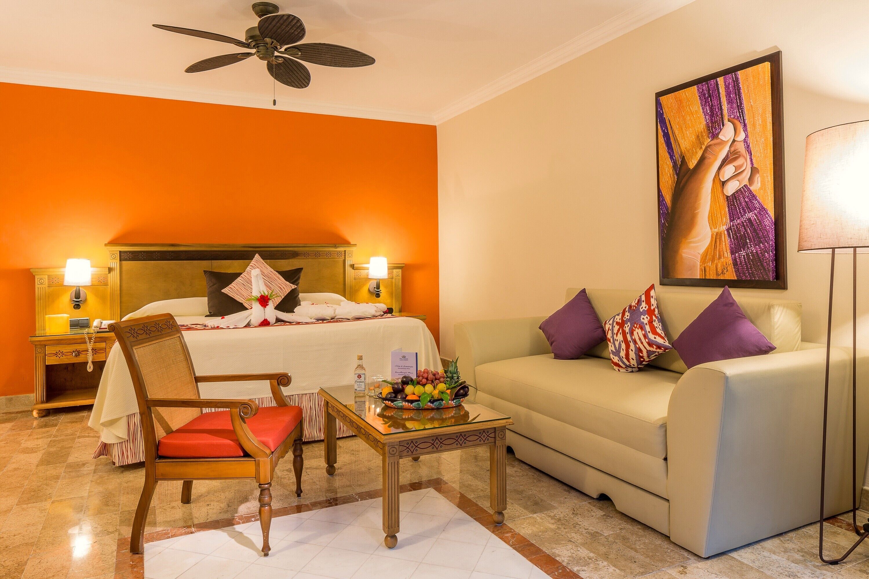 A hotel room with a bed, a sofa, a chair, a table with fruit, and a painting on the wall.