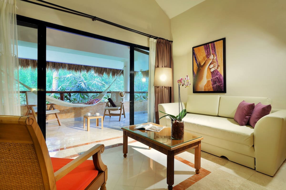 A modern living room with a glass door leading to a balcony, a white sofa, a glass coffee table, and a hammock.