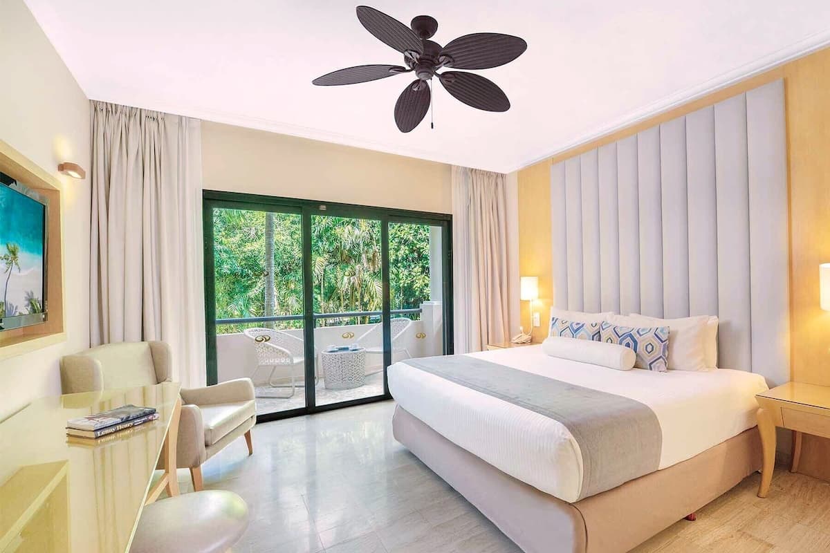 A hotel room with a bed, a desk, a chair, a ceiling fan, and a view of greenery outside.