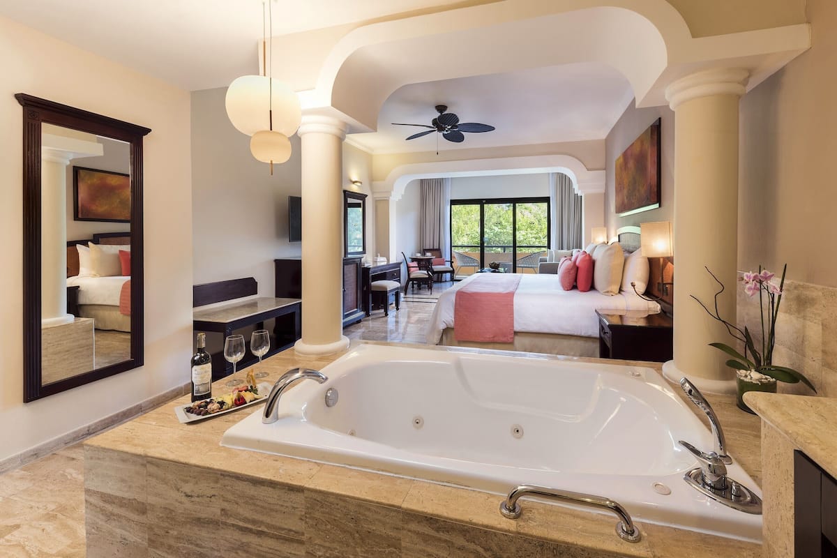 A luxurious hotel room with a large bathtub, a dining area, and a bedroom with a bed and a desk.