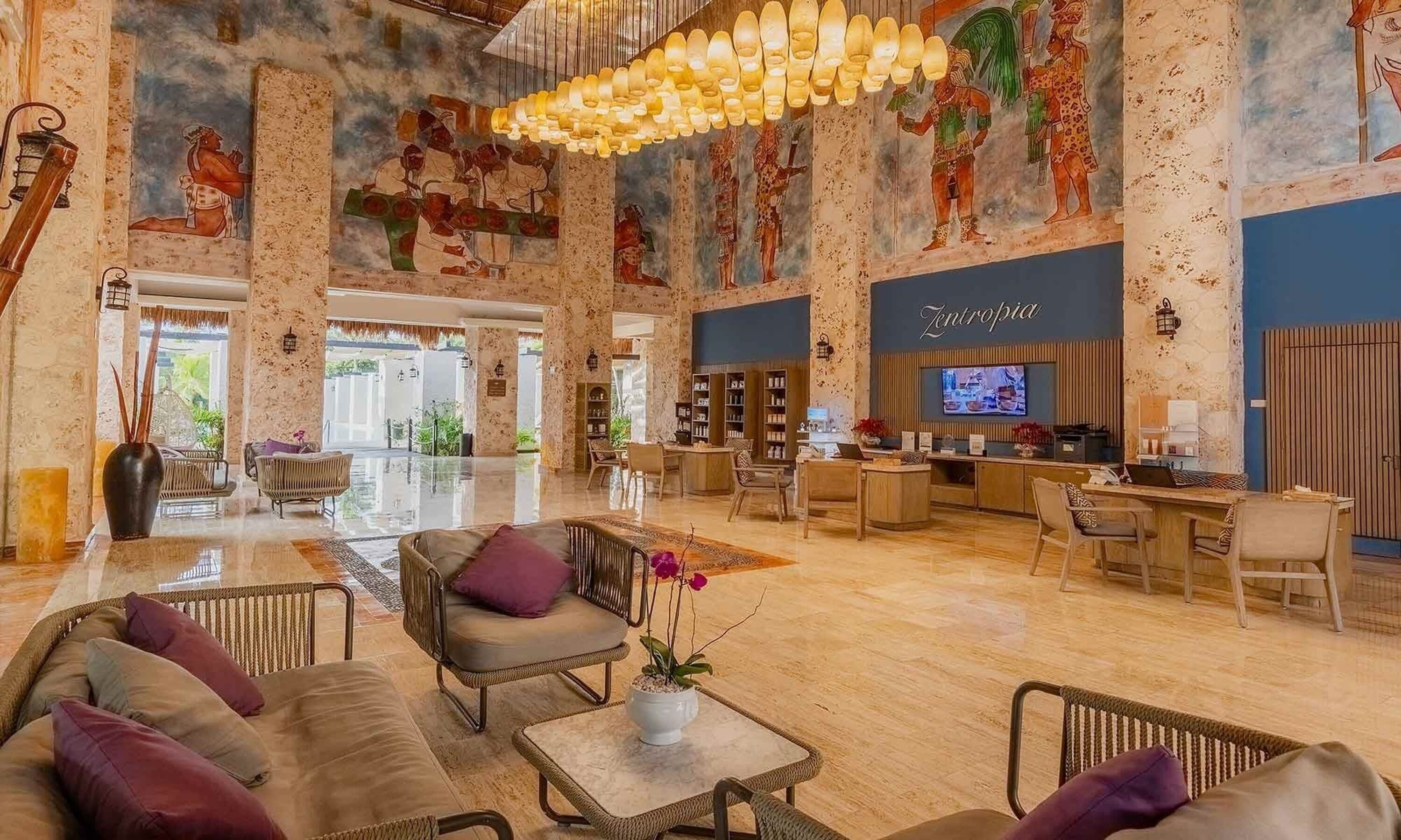 A spacious lobby with a high ceiling, a central seating area with sofas and chairs, a reception desk, and a large mural on the walls.
