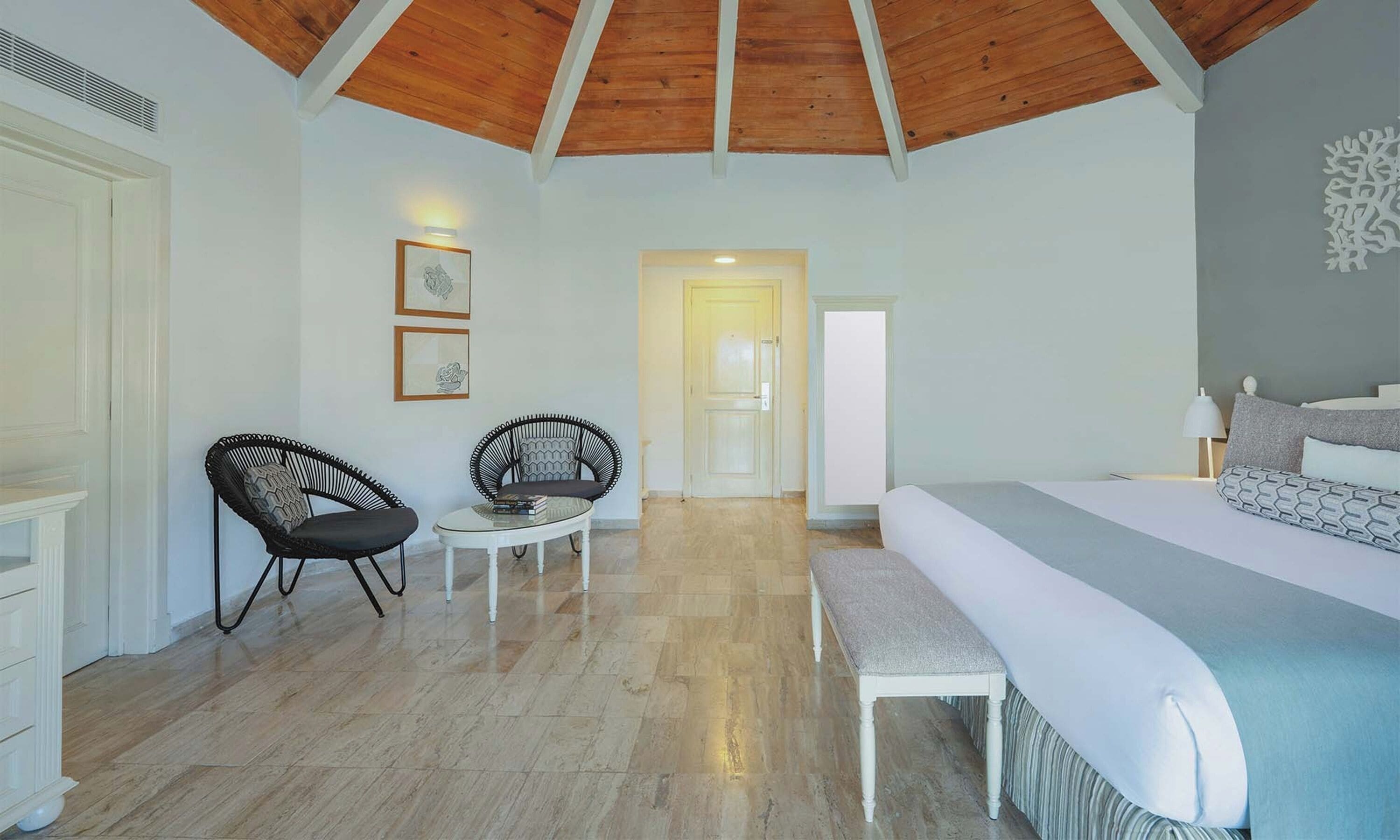 A bedroom with a bed, two chairs, a small table, and a door leading to another room.