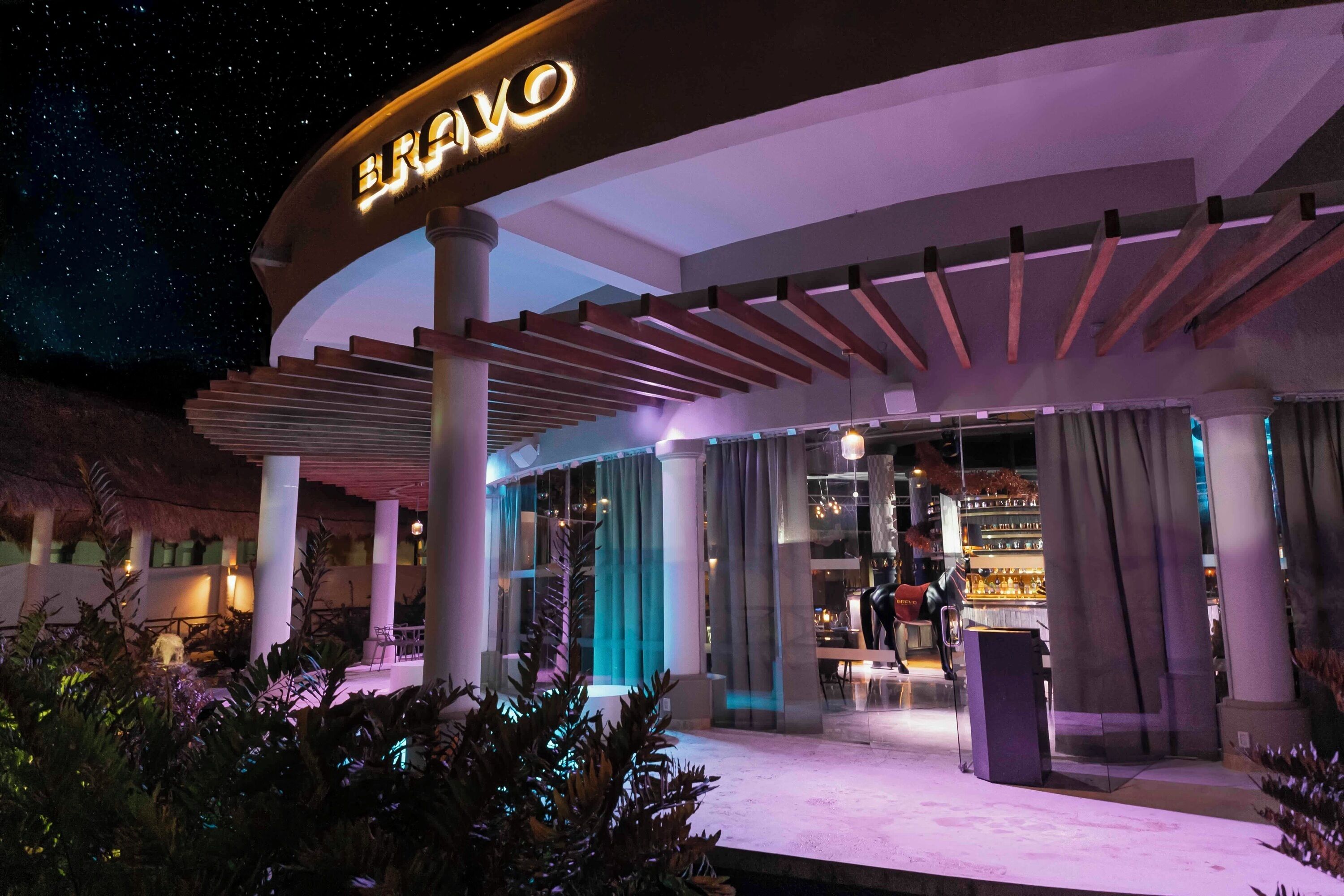 A modern hotel entrance with a prominent 'BRAVO' sign, a covered outdoor seating area, and a bar visible through the glass doors.