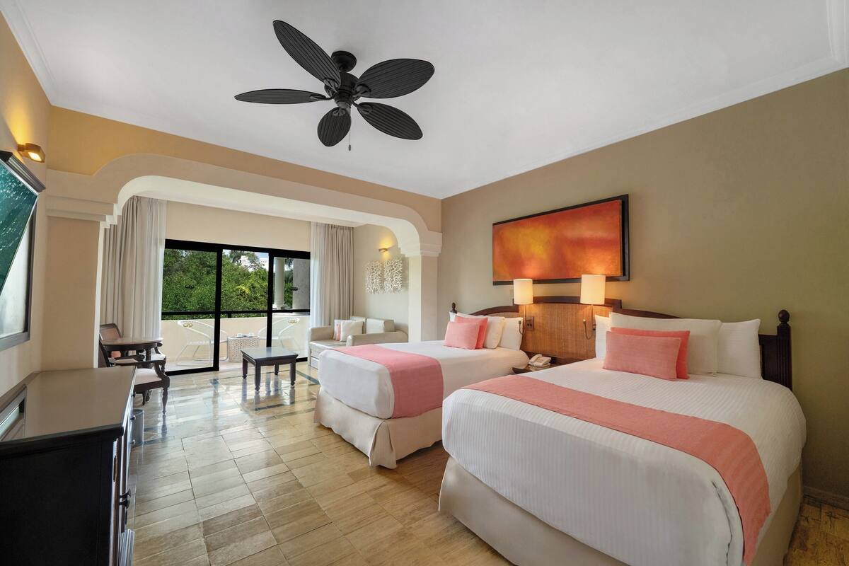 A hotel room with two beds, a ceiling fan, a desk, a chair, and a view of greenery through the window.