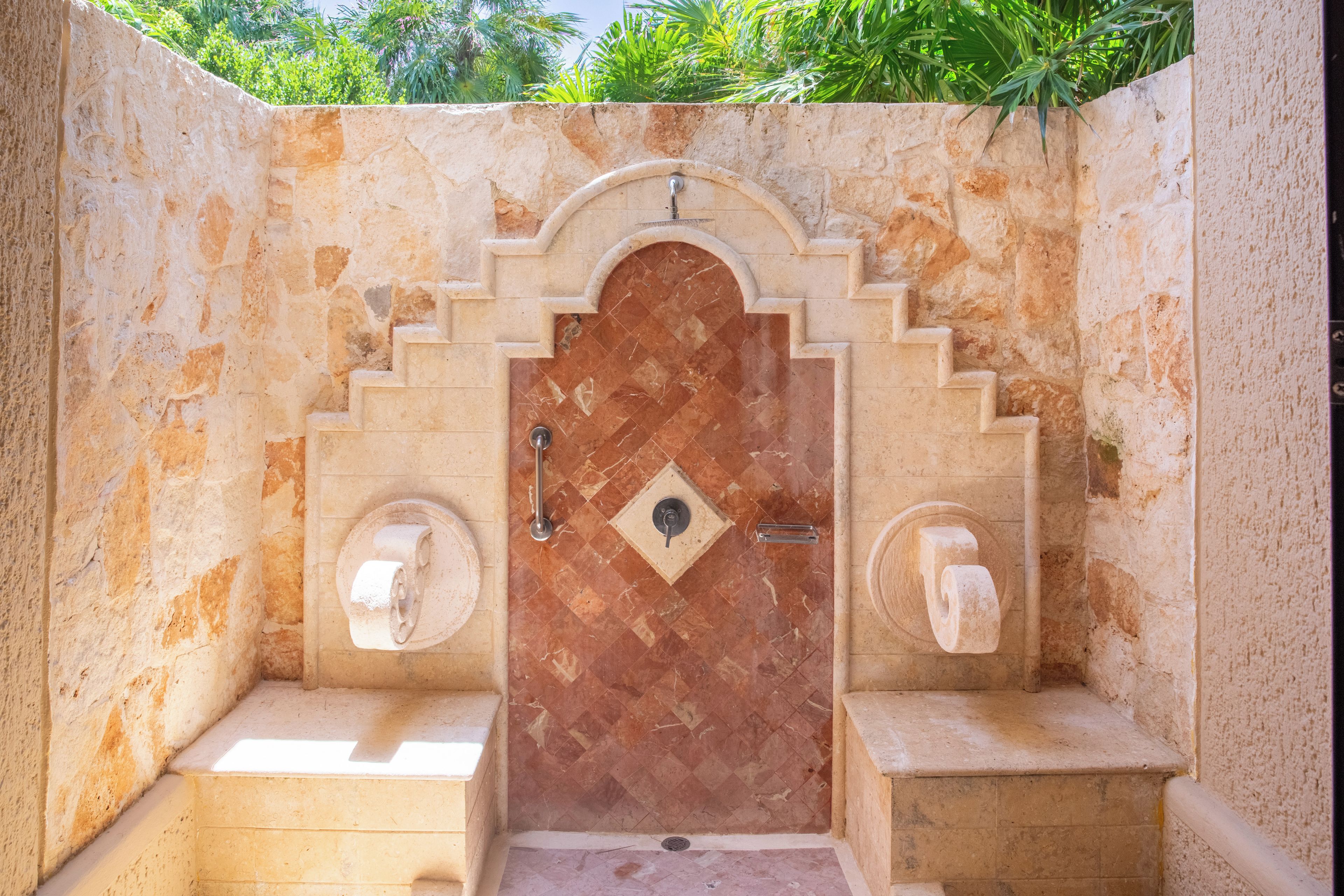 Une salle de bain aux murs en pierre, avec un sol carrelé, une porte surmontée d’une arche décorative et deux supports à papier toilette.