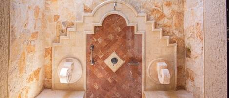 Une salle de bain aux murs en pierre, avec un sol carrelé, une porte surmontée d’une arche décorative et deux supports à papier toilette.