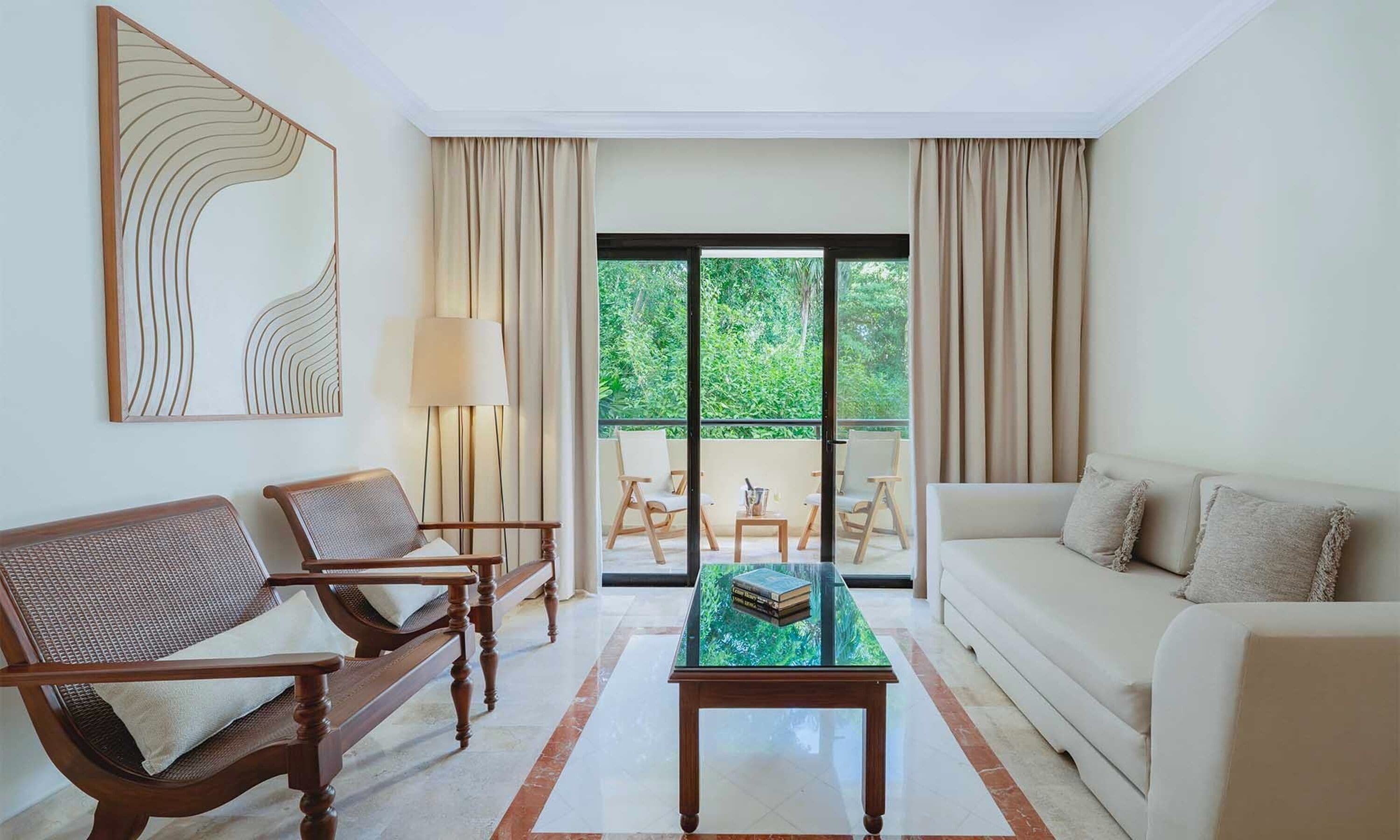 A modern living room with a sofa, two armchairs, a glass coffee table, and a large window with curtains.