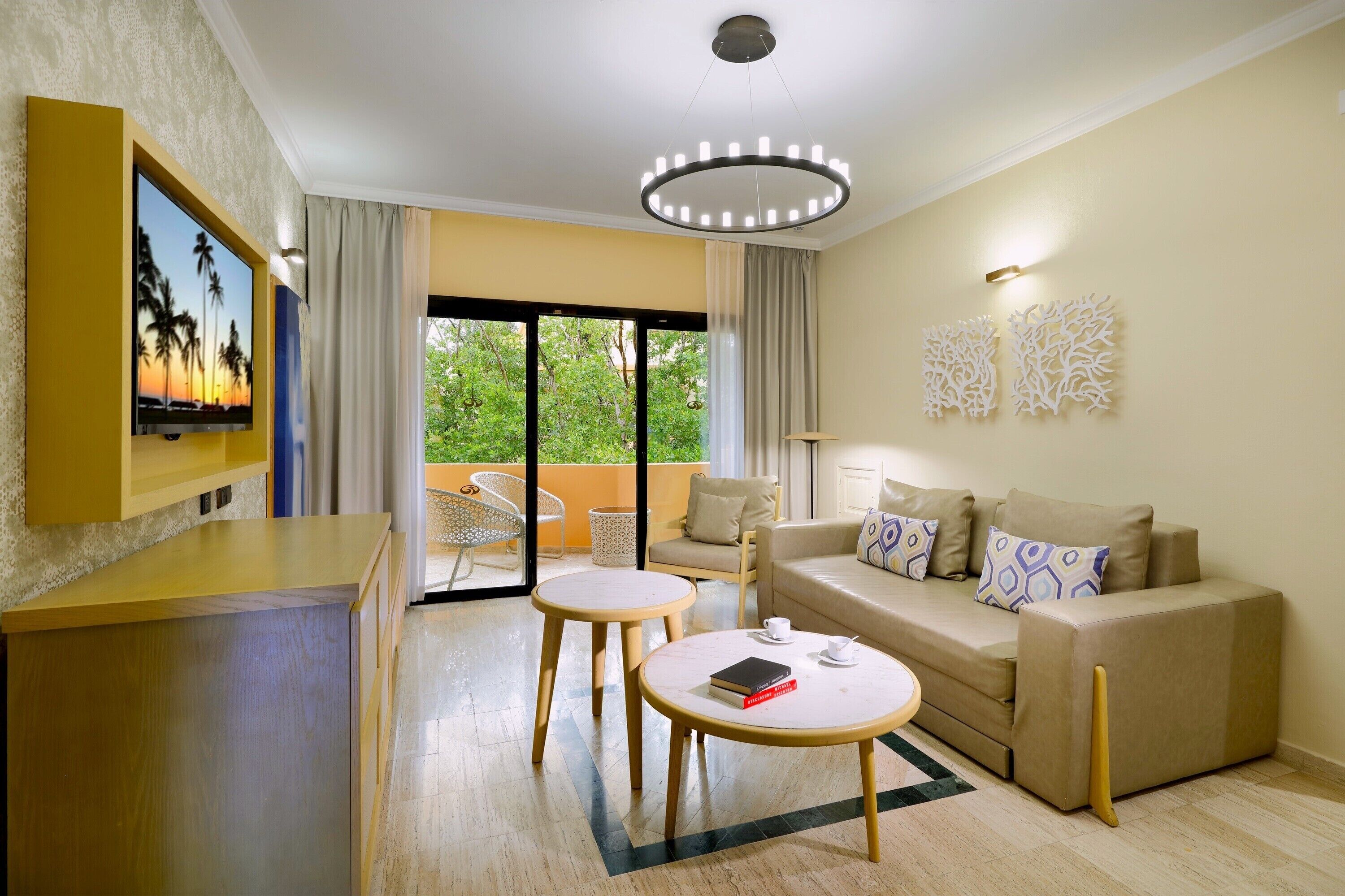 A modern hotel room with a sofa, two round tables, a TV, and a balcony with a view of greenery.
