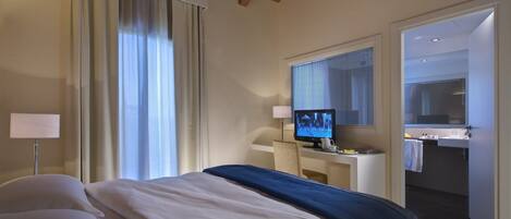 A hotel room with a bed, a desk with a computer, a window with blinds, and a bathroom visible through an open door.
