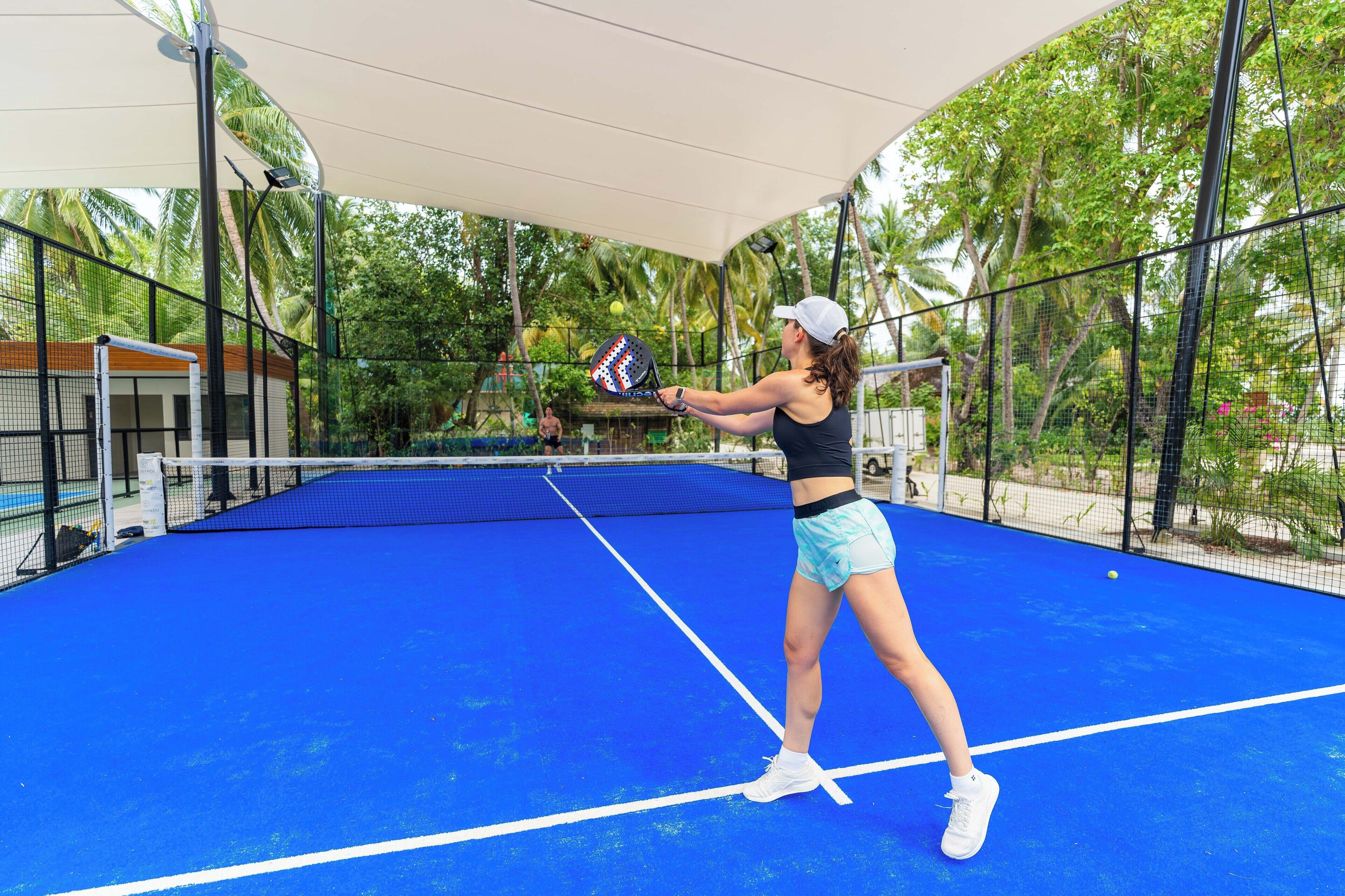 A tennis court with a net, surrounded by a fence and palm trees.
