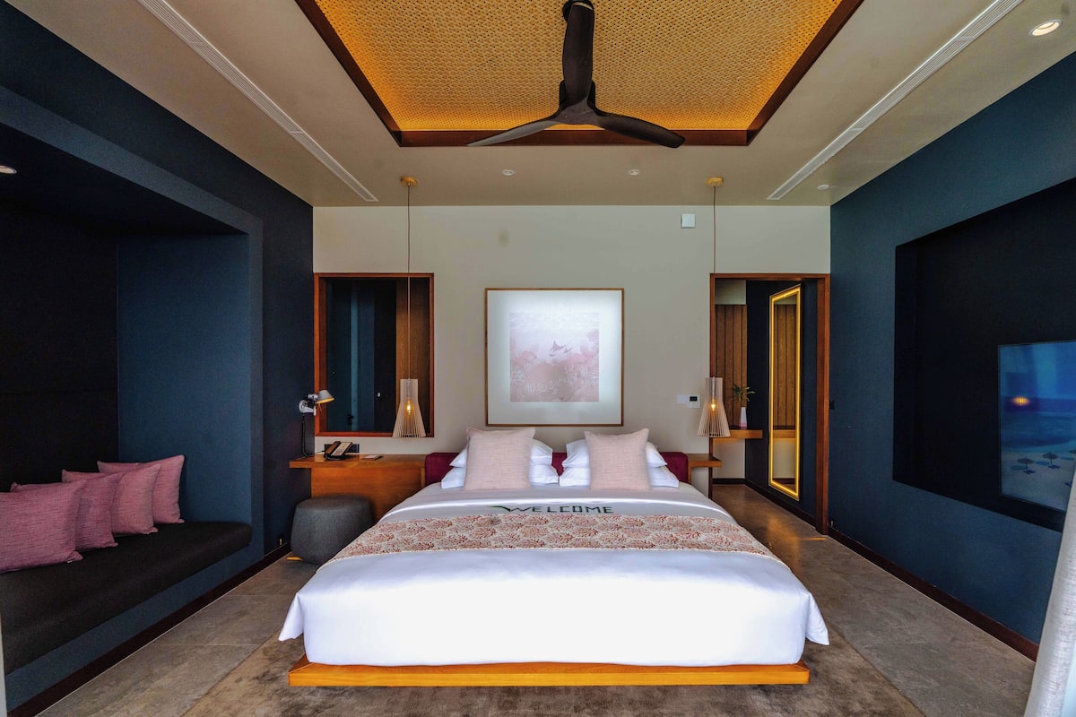 Una habitación de hotel moderna con una cama grande, un ventilador de techo, una mesita de noche, un escritorio y un ventanal amplio.