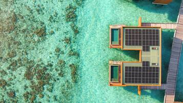An aerial view of a resort with overwater bungalows featuring solar panels, clear turquoise water, and coral formations.