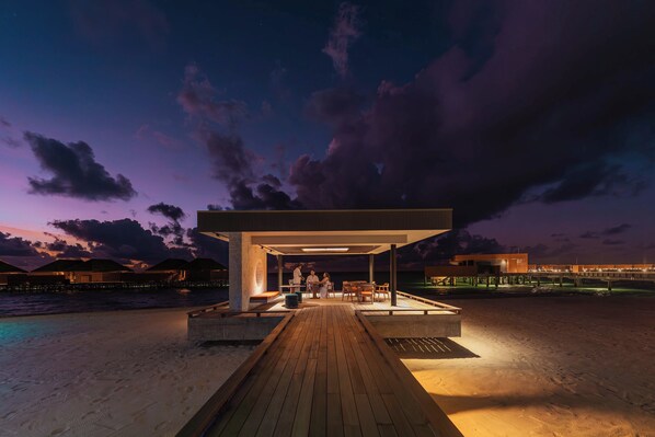 A modern outdoor lounge area with a wooden deck, seating, and a covered section with tables and chairs. - Veligandu Maldives Resort Island (Veligandu Island)