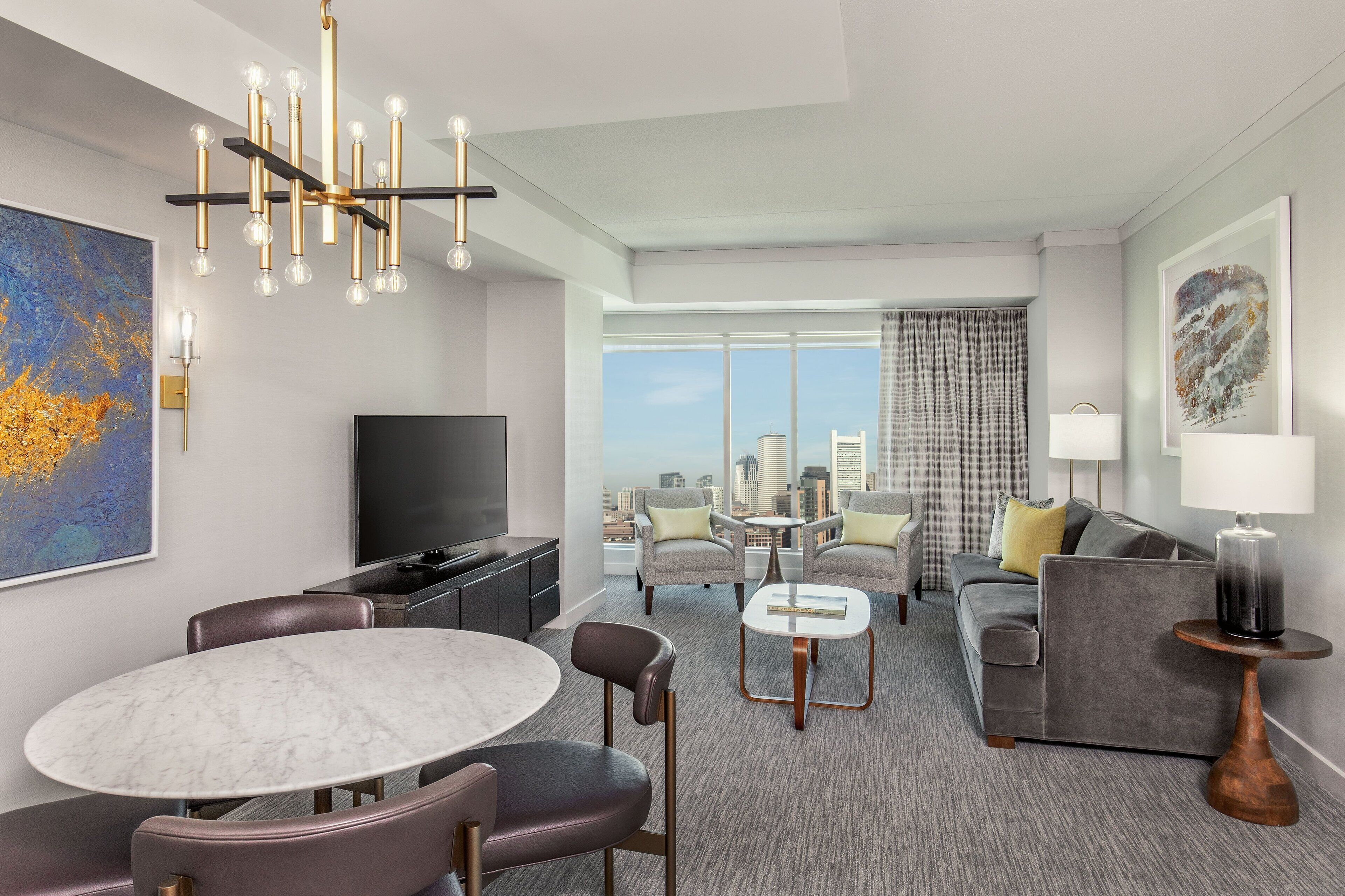 A modern hotel room with a dining area, a living space, a large window with a city view, and a flat-screen TV.